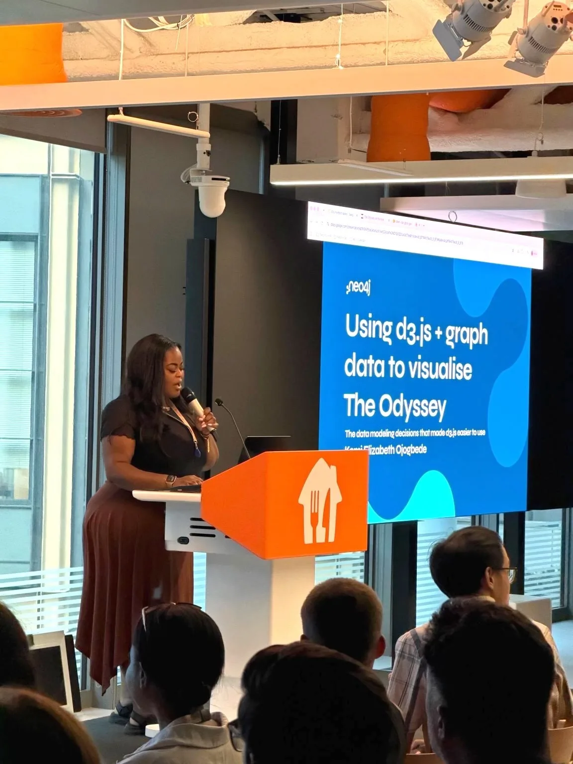 Kemi at a podium presenting a slide titled Using d3.js and graph data to visualise The Odyssey